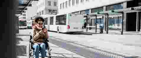 A woman using a smartphone in a wheelchair at a bus stop. A bus is in the background