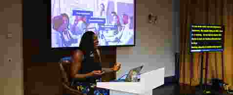 Rachel Xavier. A black woman with mid length hair sat on a stool behind a podium. She is presenting with a large presenting screen displaying "Meeting Etiquette" behind her, and there's a subtitles screen to the right. 