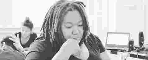 A black and white photo of a black woman with dreadlocks, looking glum and staring at a laptop. 