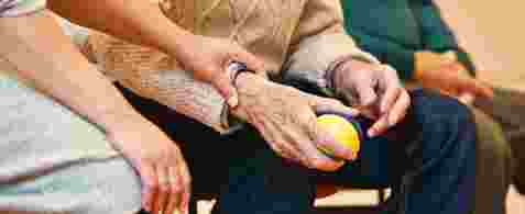 An elderly hand holding a ball. Another hand is resting on this hand with the ball. 