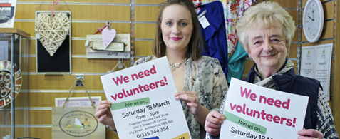 Two white women volunteering in a charity shop, holding up posters reading "We need volunteers"