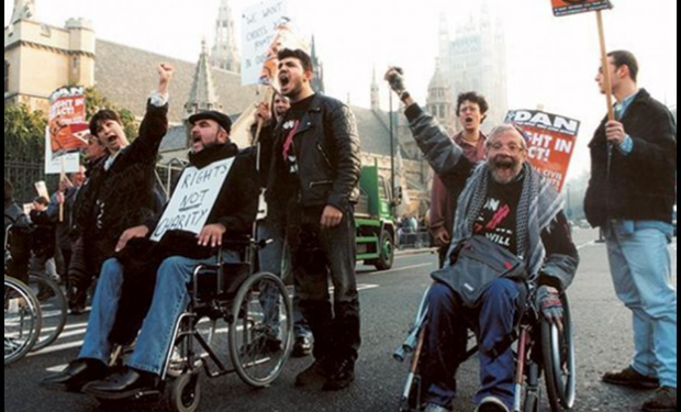 Three people in wheelchairs and six people standing are shouting and waving banners. A banner facing the camera reads "Rights Not Charity".