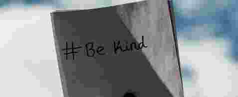 A left hand holding a note with "# Be Kind" written on it up to the sky