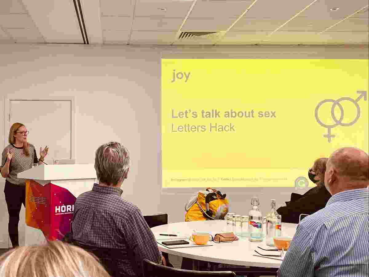 Emma Parnell gives a talk to a conference room of seated people. Behind is a slide reading 'Let's talk about sex'