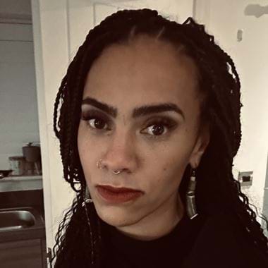 Kanika has braided hair and a nose ring stands in a room with soft lighting. She is wearing a dark top and earrings. They have a confident expression and are looking directly at the camera.