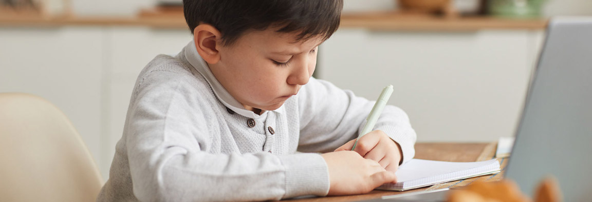 A boy is sitting and writing on a notepad. In front of him is a laptop screen.