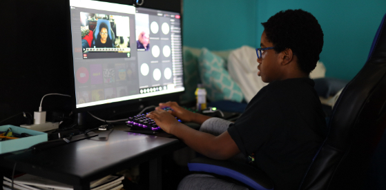 Boy wearing glasses, sitting in a semi-dark room typing on a purple qwerty keyboard while looking at a monitor screen in front of him